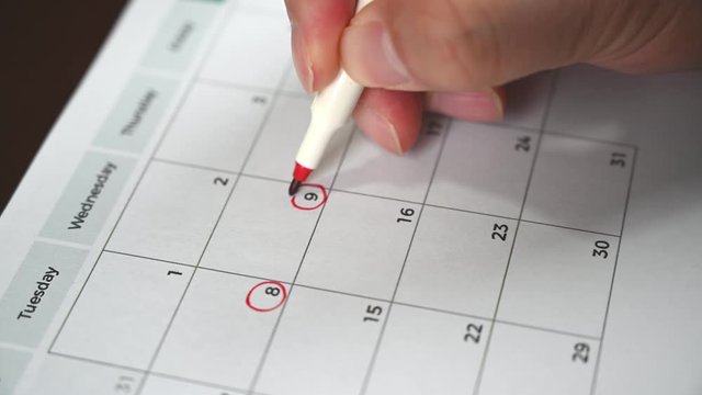 The hand of a man holding a pen in his hand and recording his schedule on a desk calendar.