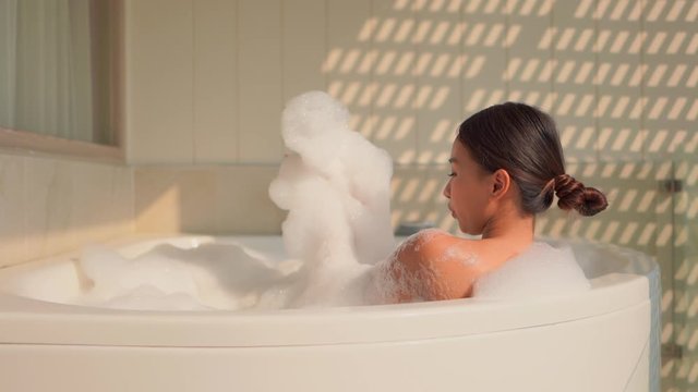 Young attractive Asian woman taking bubble bath in large tub. Over shoulder.
