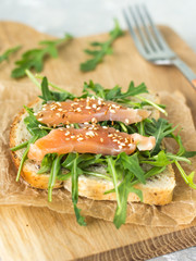 Cereal toast with arugula and trout with sesame seeds on paper and a wooden board top view