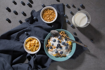 Bowl with granola and yogurt, cornflakes, blueberries and a glass of milk on a gray background