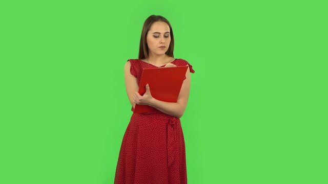 Tender girl in red dress fills papers in red folder with pensil. Green screen