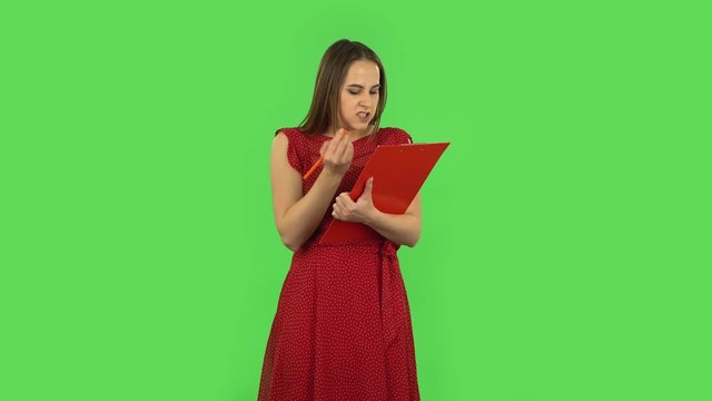 Tender girl in red dress fills papers in red folder with pensil. Green screen