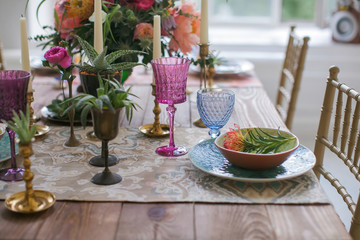 Boho chic style. Decorated wooden table with ceramic plates, cutlery, glasses for drinks, a vase with fresh flowers and candles. Golden chairs. Peonies, Succulents, orchids and Leucospermum.