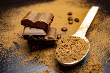 Pieces of porous chocolate, a wooden spoon with cocoa, coffee beans on a black background