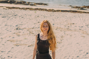 Portrait of young smiling blond girl in black blouse and skirt  walking in the day on the white beach near the sea