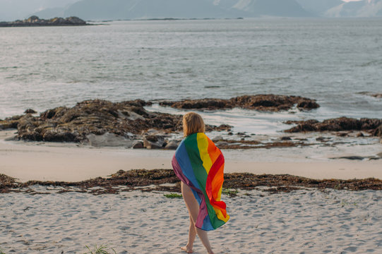 Young naked blond girl covered with LGBTQI flag walking in the day on the white beach near the sea