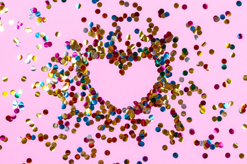 Heart shape made of golden colorful confetti splash on pink background. Festive backdrop of sparkles for birthday, valentine's day, carnival