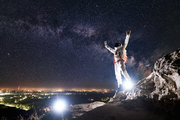 Spaceman raising arms while standing on rocky mountain with fantastic starry sky on background. Astronaut wearing white space suit and helmet. Concept of space travel, milky way and nighttime.