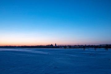 Snowy plain under the sunset sky. Bell tower small town skyline. 