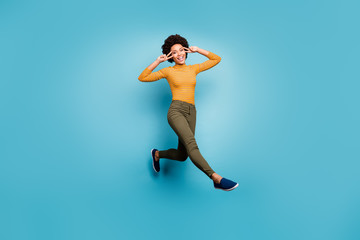 Full size photo of cheerful afro american girl have fall weekends holidays jump make v-signs wear yellow green stylish clothing isolated over blue color background