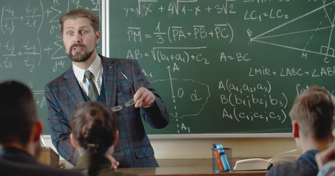 Handsome math teacher in suit holding eyeglasses and explaining lecture to students