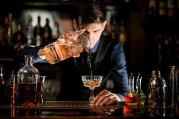 Barman holding glass of cocktail and pours it into wineglass.