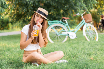 Obraz premium Beautiful young woman sitting on grass and drink orange juice on vintage bicycle background in summer park.
