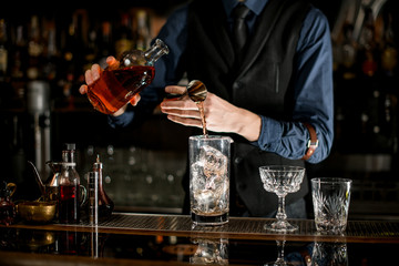 Bartender measures the ingredients for cocktail from bottle with brown alcohol drink.