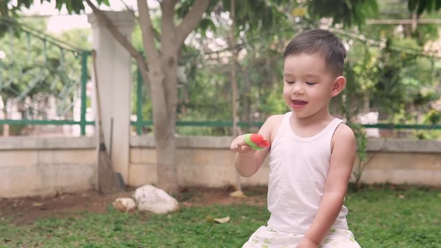 Slow motion video of a handsome asian kid eating a delicious watermelon red ice cream in the backyard