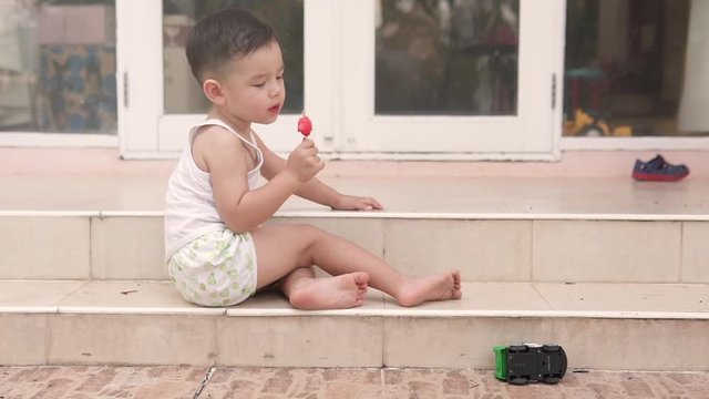 Slow motion video of a handsome Asian kid from the side eating a posicle ice cream sitting on the stairs of a house in summer