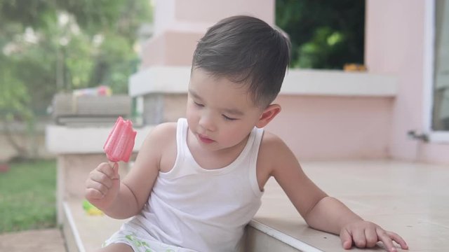 Slow motion video of a handsome Asian boy sitting on the stairs outside of the house happy singing a song while holding a red strawberry ice cream
