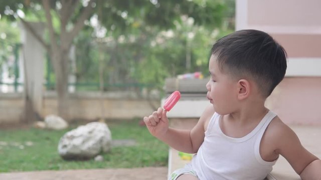 Slow Motion video of Asian boy eating strawberry ice cream sitting on the stairs outside of the house in the yard and looking around in a summer afternoon