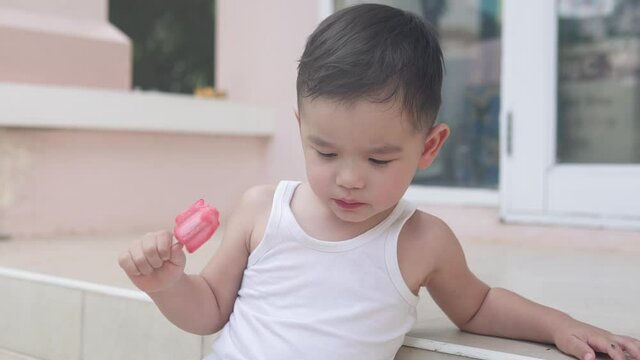 Asian toddler boy sitting on the stairs outside of a house focused on eating a red strawberry ice cream popsicle with a serious face