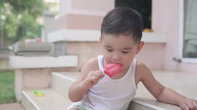 Slow motion Shallow depth of field video of a cute Asian boy calmly eating a strawberry popsicle ice cream  sitting on the stairs outside of his house