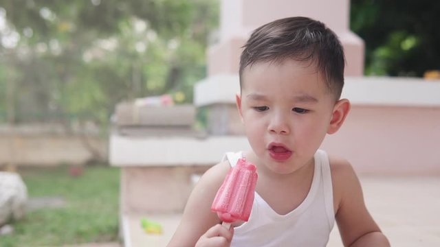 Shallow depth of field video of a cute Asian boy eating and enjoying a strawberry red  popsicle ice cream In his house backyard