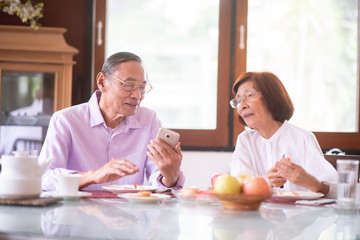 Fototapeta na wymiar Asian elderly couple using smart phone for social media and eating fruits and talking at home.retirement people and technology concept