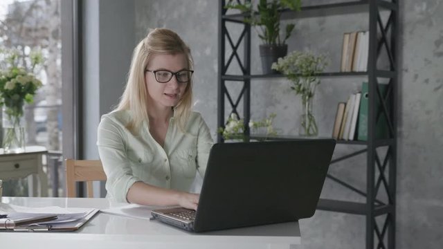 business online, happy attractive girl freelancer works at a laptop computer in the office with beautiful fresh flowers interior then shrugs hands to side rejoicing at job done and career success