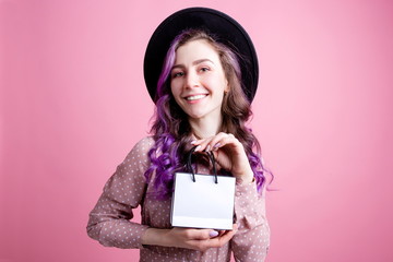 Surprised beautiful girl with purple hair standing on a pink background holding a gift bag