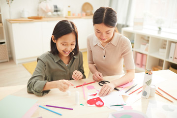 Young Asian woman and her teen sister creating handmade Mothers Day holiday card with hearts,...