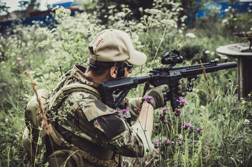 A military man or airsoft player in a camouflage suit sits in the grass and aims from an automatic...