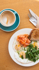 Tasty healthy breakfast: fried eggs, salmon, arugula and croutons on a white plate and  a mug of cappuccino on a wooden table.
