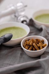 Spring detox broccoli green cream soup in white plates, bowl with crackers and stylish black cutlery on grey background. 