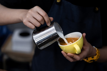 Close-up of barista hand holding and pouring hot milk for prepare latte art on a yellow cup of cappuccino coffee.