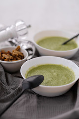 Spring detox broccoli green cream soup in white plates, bowl with crackers and stylish black cutlery on grey background. 
