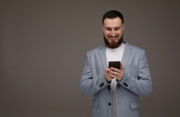 Handsome bearded model is posing with phone in hands over grey background.