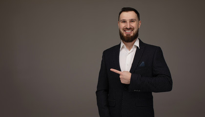 Close up portrait of young successful brunette stock-market broker guy on grey background, he is smiling, wearing suit and is pointing on a copy space with his finger.