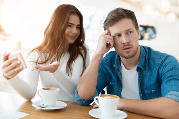 young woman drinks coffee with her husband, she shows him photoes on the phone while he turned around looking tired, digital influence concept