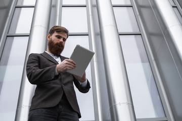 Modern Young bearded Business man working with a digital tablet. Young hipster businessman holding tablet in hands outdoor. Working online with a tablet while standing outside on an office building.