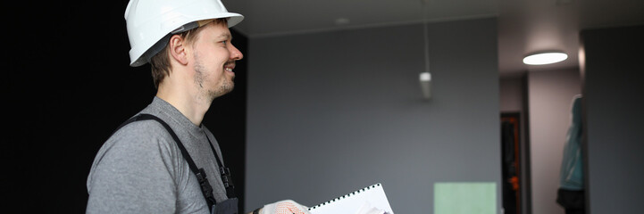 Young smiling repairman standing in empty flat and holding construction plan in hands