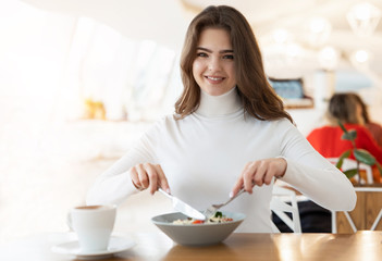 young positive beautiful woman eats salad with knife and fork in cafe during lunch time, food and drink concept