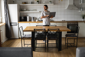 Freelancer working from home on the kitchen and using laptop. Bearded man working with a laptop and reading good news. Handsome successful self entrepreneur standing with laptop and working at his