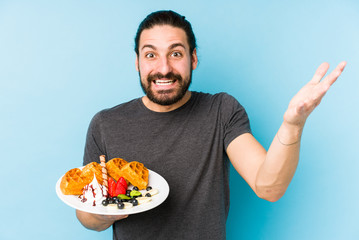 Young caucasian man eating a waffle dessert isolated receiving a pleasant surprise, excited and...