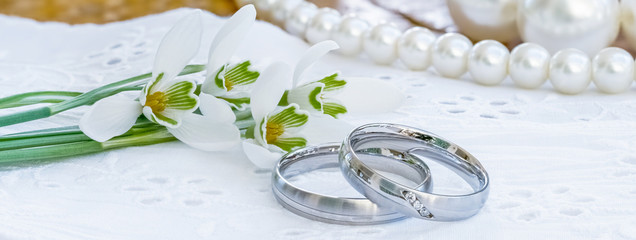 Silberne Eheringe liegen neben Schneeglöckchen und weißen Perlen auf einem Holztisch