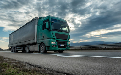 Green semi trailer truck driving on a highway with dramatic sunset sky in the background....