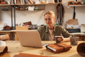 Fototapeta premium Horizontal portrait of young Caucasian woman sitting at wooden table in her workshop using laptop