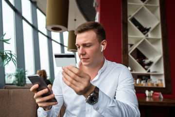Young businessman wearing a white fashion shirt paying via mobile cell phone and credit card in a cafe with a laptop. Freelance and selfemployment concept. Distance dream job.
