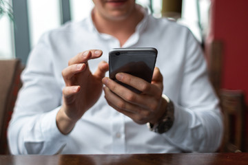 Closeup of a young businessman hands wearing a white fashion shirt chatting on a mobile cell phone in a cafe with a laptop. Freelance and selfemployment concept. Distance dream job.
