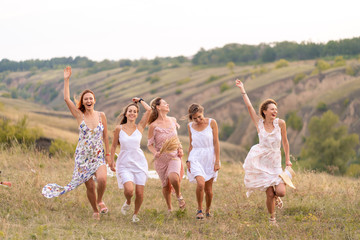 Fototapeta premium The company of cheerful female friends have a great time together on a picnic in a picturesque place overlooking the green hills. Girls in white dresses dancing in the field