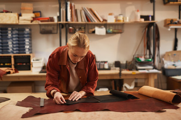 Fototapeta na wymiar Horizontal shot of young Caucasian woman wearing casual outfit working in leather craft studio