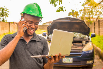 young african man, a professional mechanic, using a laptop to fix and detect fault in a car, making a phone call